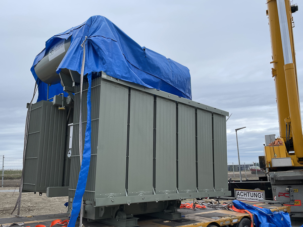 En kran, der transporterer en stor, cylindrisk transformer dækket af en blå presenning. Der er et advarselsskilt på siden af lastbilen.