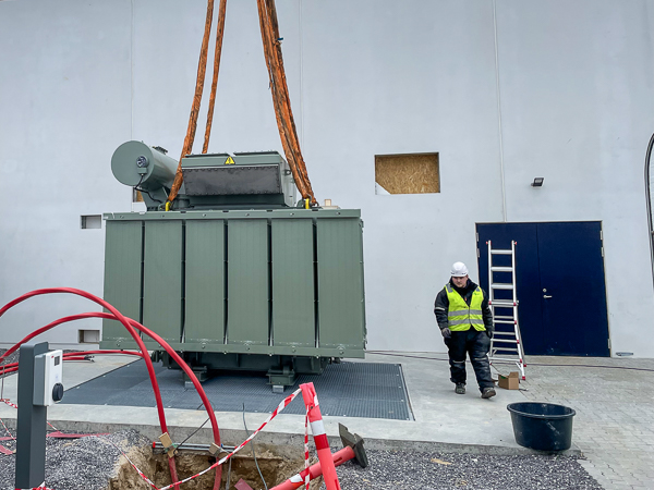 Large transformer being mounted on a foundation. The transformer is handled by a large crane.