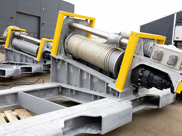 A close-up of a yellow winch with a dynema cable wrapped around its drum. The winch sits on wooden pallets in front of a building.