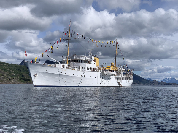 Kongeskibet "Norge" sejler i en malerisk fjord under en skyet himmel, dekoreret med flag.