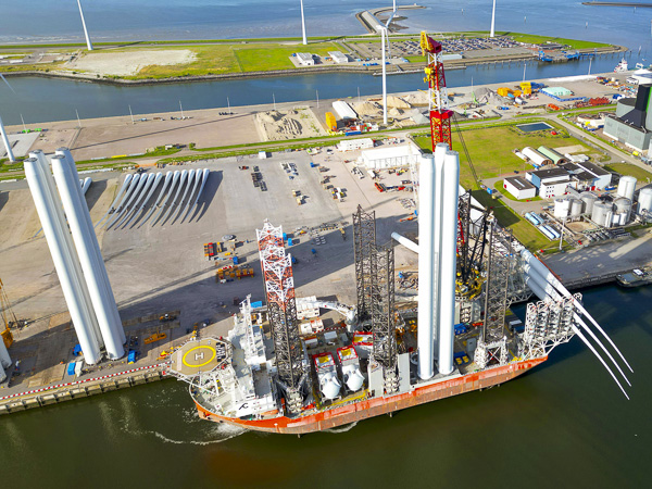 Aerial view of a port with a ship carrying parts of a wind turbine, including tower sections and blades, ready for transportation. Several wind turbines are visible in the background.