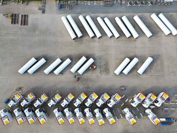 Aerial view of a storage area with multiple wind turbine components, including tower sections and nacelles, arranged neatly for transportation.