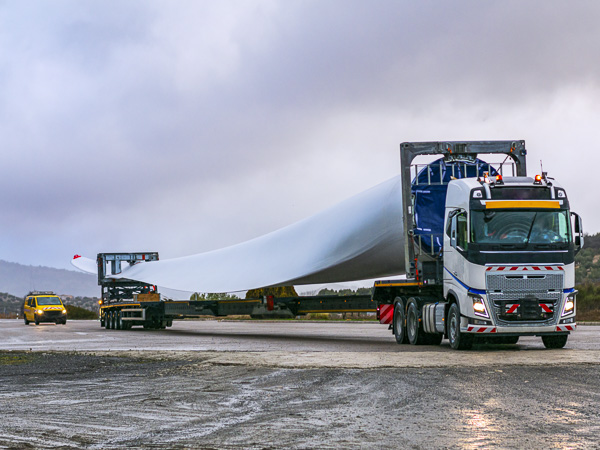 A truck transporting a single wind turbine blade on an extended trailer. An escort vehicle is visible, indicating a controlled transportation process.