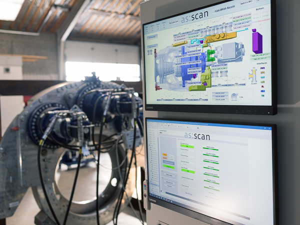 Two monitors displaying AS SCAN's software interface for managing wind turbine components, with a large industrial gearbox assembly in the background in a workshop setting