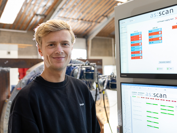 En mand i en AS:SCAN-trøje smiler ved siden af en Turner Gear Test Board-skærm, der viser testresultater i et industriværksted.