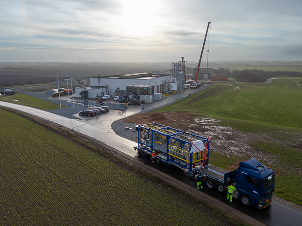 Truck with a large process module arrives at the facility, with a crane and buildings in the background.