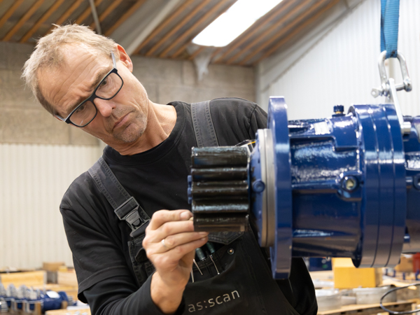 En AS SCAN-medarbejder iført sorte overalls og en sort T-shirt, inspicerer et stort sort tandhjul foran en nymalede blå maskindel, der er ophængt i en løfteanordning.