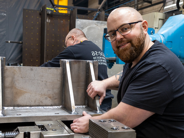 En AS SCAN-medarbejder med skæg og briller smiler til kameraet, mens han arbejder på en stor, svejset metalstruktur på et værksted. En anden medarbejder i en AS SCAN-sweatshirt er synlig i baggrunden.
