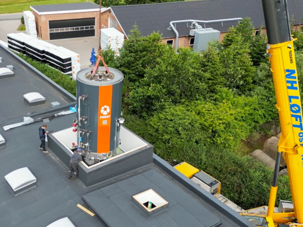 Luftfoto af to teknikere, der overvåger den kranassisterede installation af en stor, cylindrisk lagertank (mærket 'Parat' med orange striber) på taget af en industribygning. En gul 'NH LØFT'-kran er synlig i højre side.