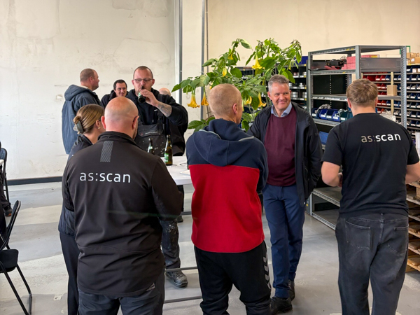 Employees standing and talking at a high table in the workshop, with others seated in the background during the informal gathering.